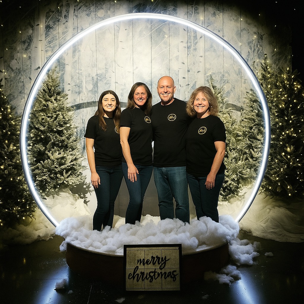 Cztery osoby w czarnych koszulkach z logo stoją w dekoracji świątecznej z napisem 'Merry Christmas' i choinkami, oświetlone neonowym okręgiem.