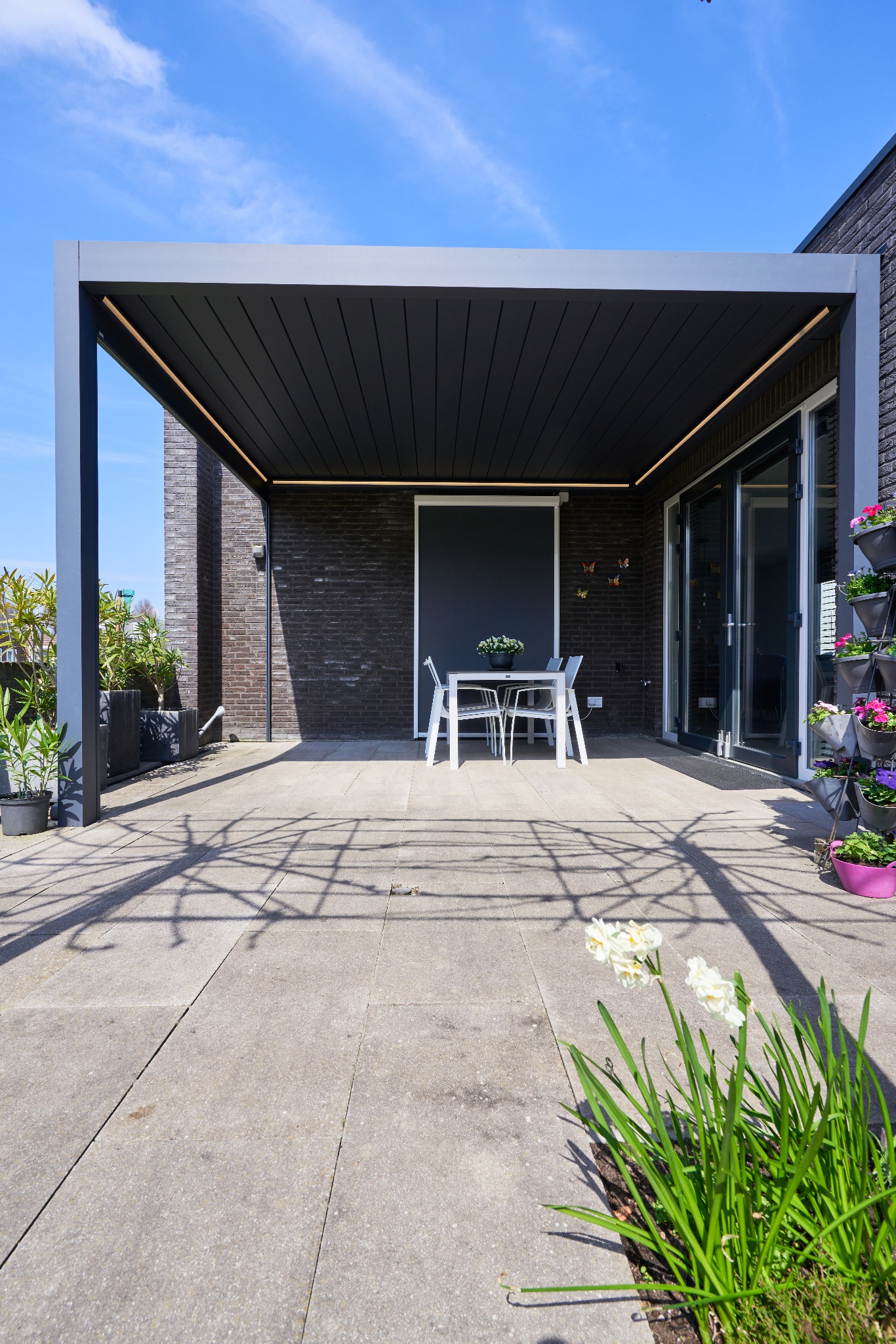 Nowoczesna, grafitowa konstrukcja z oświetleniem LED, zacieniająca taras z białym stołem i krzesłami. W tle ceglany budynek i kwiaty w donicach.