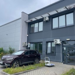 Ventico Karol Kuźniak - Montaż klimatyzacji na elewacji budynku. Pracownik instaluje jednostkę zewnętrzną. Widoczny samochód i wózek transportowy. Szara elewacja i kostka brukowa.