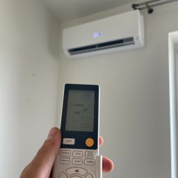 Ventico Karol Kuźniak - Biały klimatyzator ścienny z wyświetlaczem 19°C, sterowany pilotem w dłoni. Nowoczesny design, jasne wnętrze. Ustawiona temperatura na pilocie.