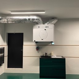 Ventico Karol Kuźniak - Biała centrala rekuperacyjna z srebrnymi przewodami wentylacyjnymi zamontowana w pomieszczeniu z zieloną podłogą i ciemnozieloną szafką z umywalką.