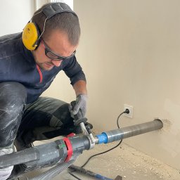 Ventico Karol Kuźniak - Pracownik w okularach i słuchawkach wierci otwór w ścianie za pomocą wiertnicy z odsysaniem pyłu. Widoczne narzędzia i kabel zasilający. Praca w trakcie wiercenia.