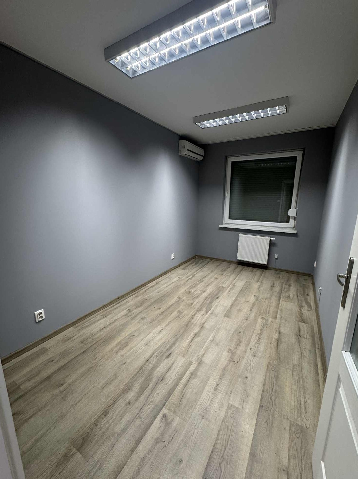 Wnętrze biura po odświeżeniu: szare ściany, jasna podłoga imitująca drewno, białe okno z grzejnikiem i nowoczesne oświetlenie sufitowe.