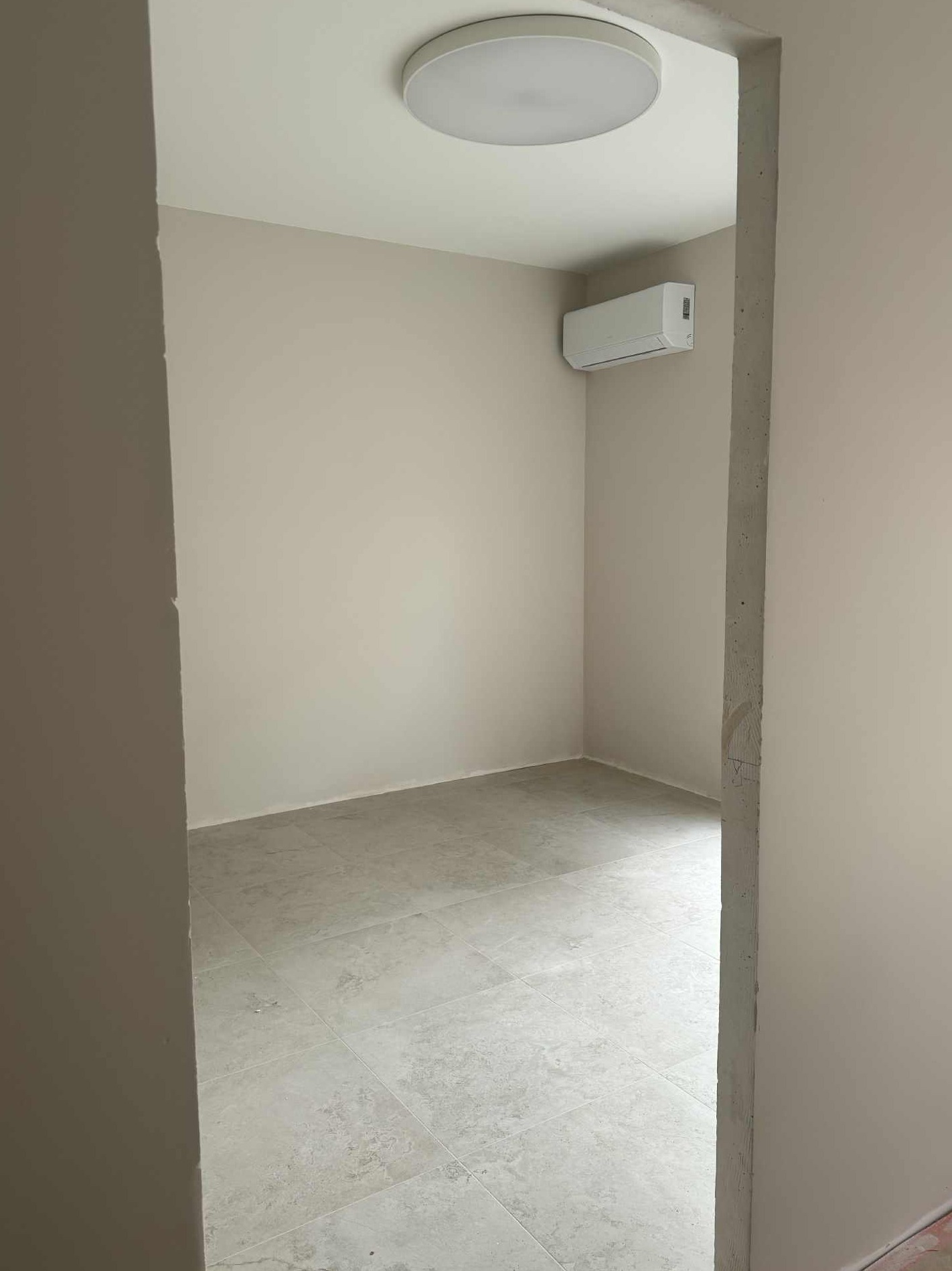 Jasne, świeżo pomalowane wnętrze z klimatyzacją i płytkami podłogowymi. Widok zza framugi, neutralna kolorystyka ścian i sufitu, okrągła lampa sufitowa.