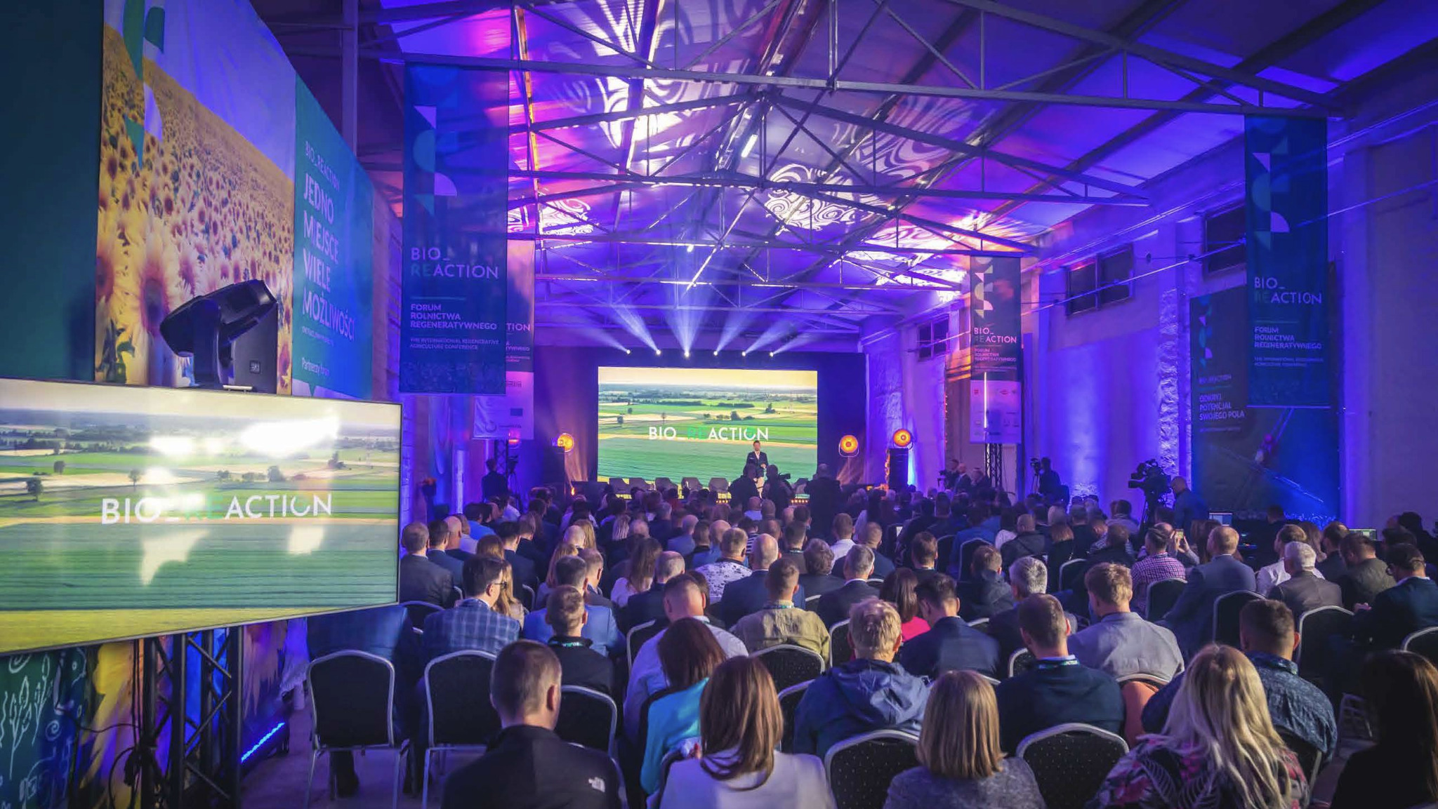 Konferencja w dużej sali z widownią, scena z ekranem i prelegentem, oświetlenie punktowe, banery informacyjne. Widok z tyłu sali, skupienie na scenie i widzach.