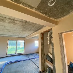 Expert-Bud Sebastian Śmigiel - Surowy strop betonowy z instalacją, ściany po wstępnym malowaniu, rury ogrzewania podłogowego na posadzce. Widok wnętrza w trakcie prac wykończeniowych.