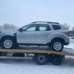 Pomoc w Trasie - pomoc drogowa, laweta, transport, awaryjne uruchamianie Częstochowa - Srebrny SUV na żółtej lawecie w zimowej scenerii. Widoczny numer telefonu na kabinie lawety. Auto zabezpieczone pasami. Tło: parking i drzewa.