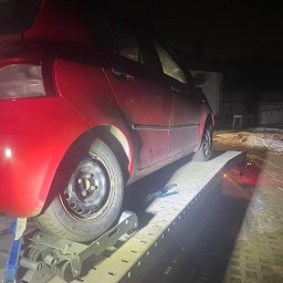 Pomoc w Trasie - pomoc drogowa, laweta, transport, awaryjne uruchamianie Częstochowa - Czerwony samochód osobowy z uszkodzonym kołem, zabezpieczony na platformie lawety w nocnych warunkach. Widoczne detale mocowania i oświetlenie.