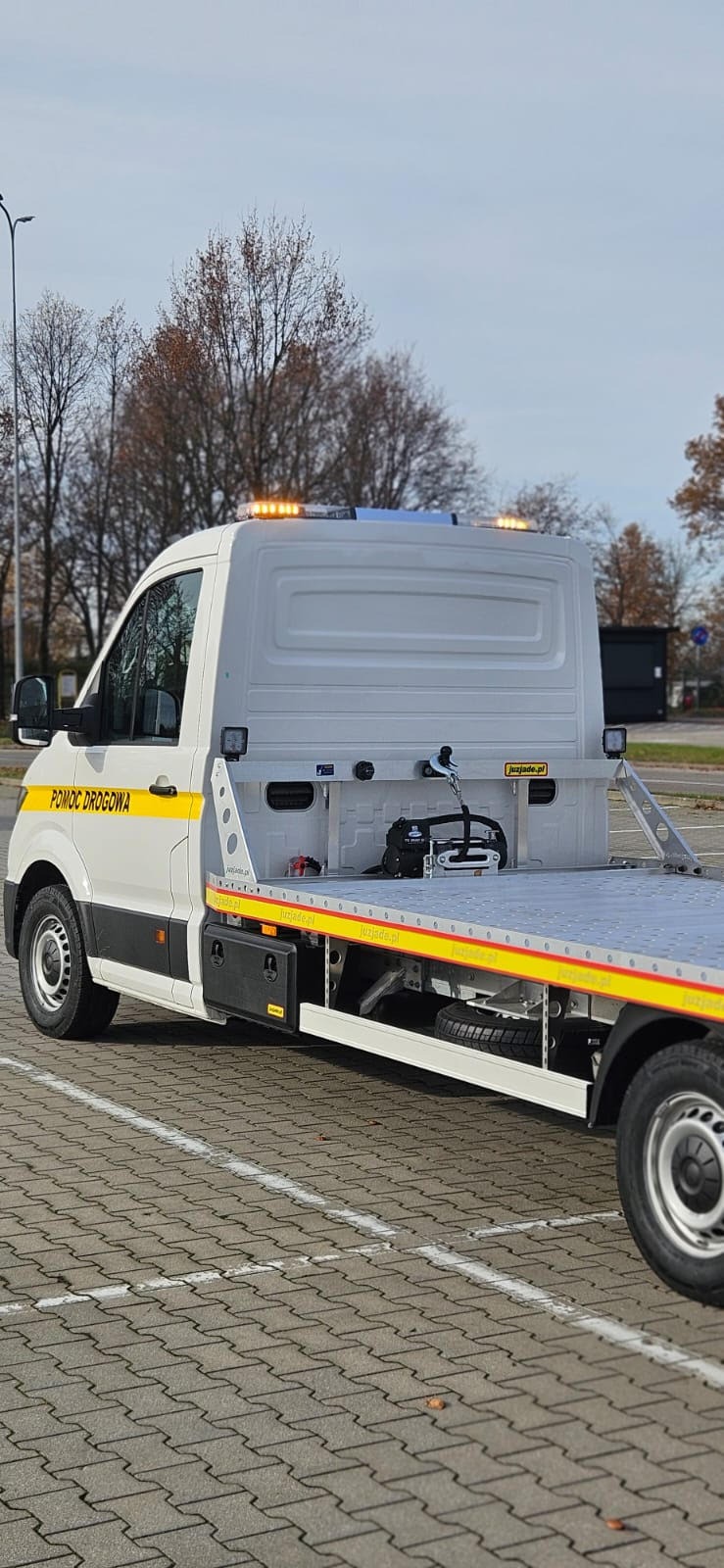 Biała laweta z napisem 'Pomoc Drogowa' na parkingu z kostki brukowej. Widoczne światła ostrzegawcze i platforma załadunkowa. Jesienne drzewa w tle.