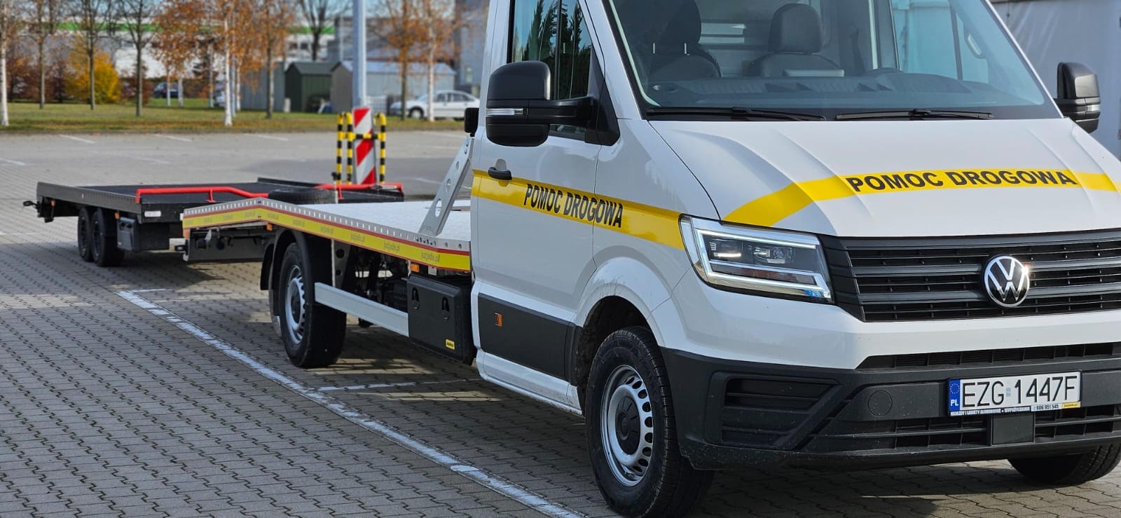 Biała laweta Volkswagen z żółtym pasem i napisem 'Pomoc Drogowa' na parkingu z kostki, jesienne drzewa w tle. Widoczny fragment tablicy rejestracyjnej.