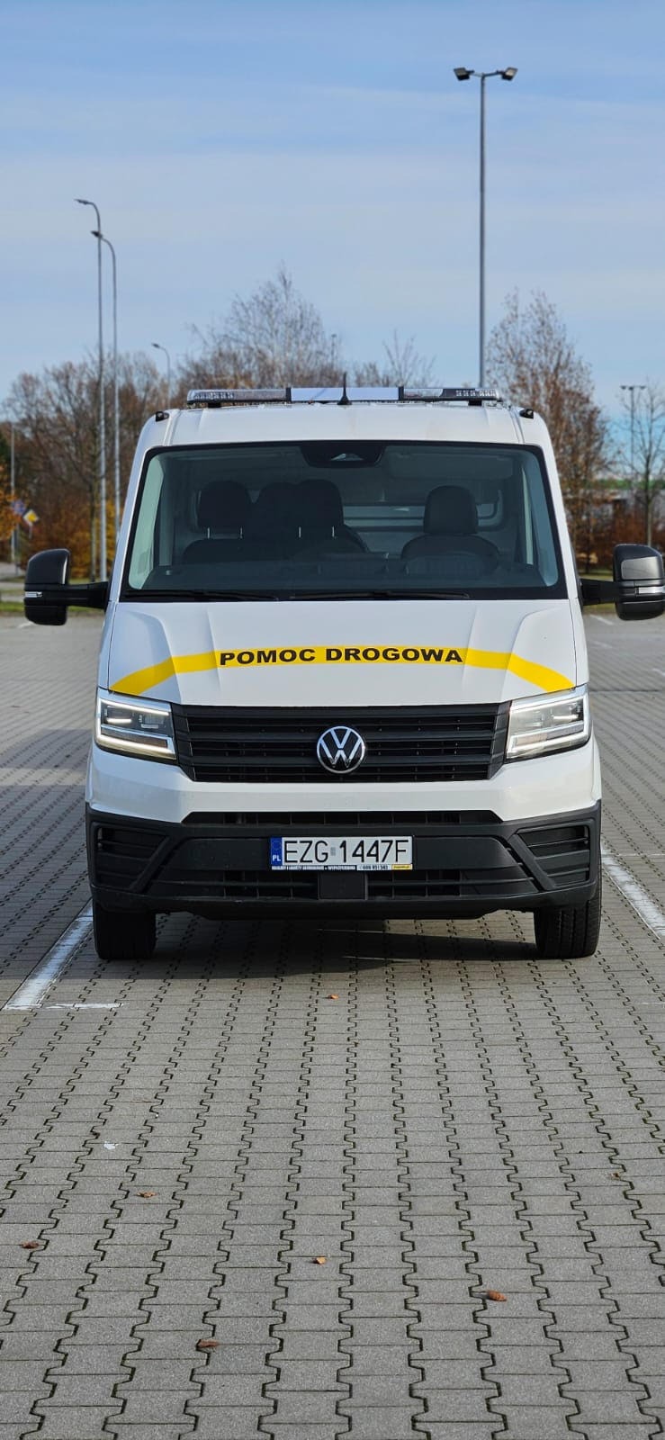 Biały bus pomocy drogowej Volkswagen z żółtym pasem i napisem 'POMOC DROGOWA' na parkingu z kostki, widok z przodu, tablica rejestracyjna widoczna.