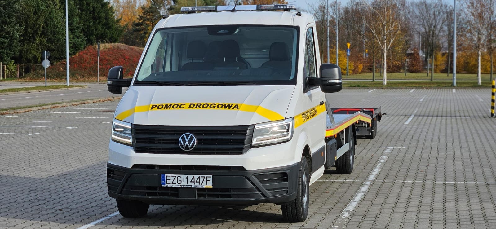 Biała laweta Volkswagen z żółtym pasem 'Pomoc Drogowa' na parkingu z kostki, jesienne drzewa w tle. Tablica rejestracyjna widoczna.