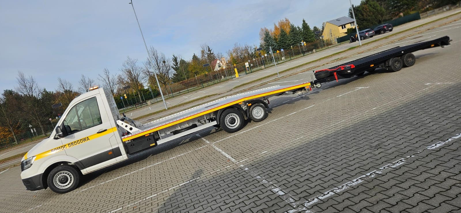 Biała laweta z żółtymi pasami 'Pomoc Drogowa' na parkingu, ciągnąca czarną przyczepę. Jesienne drzewa w tle, błękitne niebo.