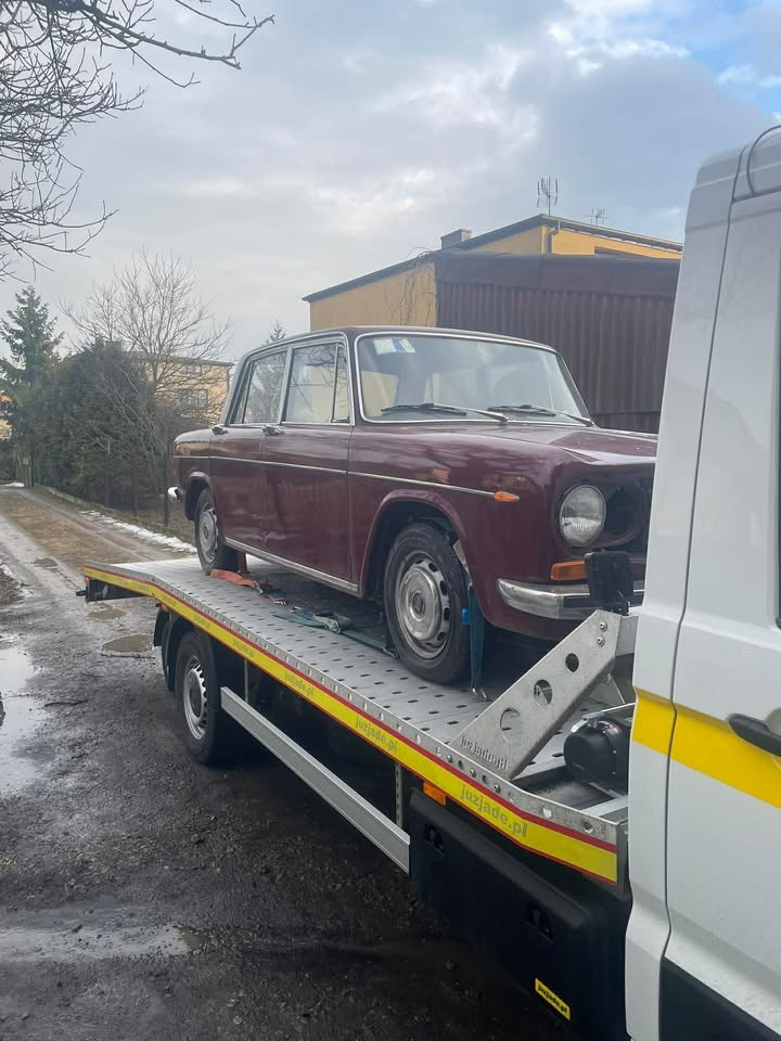 Bordowy, zabytkowy samochód osobowy zabezpieczony pasami na platformie lawety. Widoczne budynki w tle i fragment pojazdu lawety z żółtymi pasami.