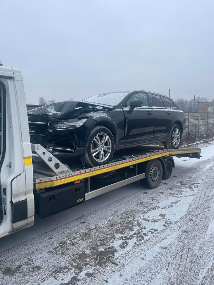 Czarny, uszkodzony samochód osobowy na żółtej platformie lawety w zimowej scenerii. Widoczne zniszczenia przodu pojazdu i śnieg na karoserii.