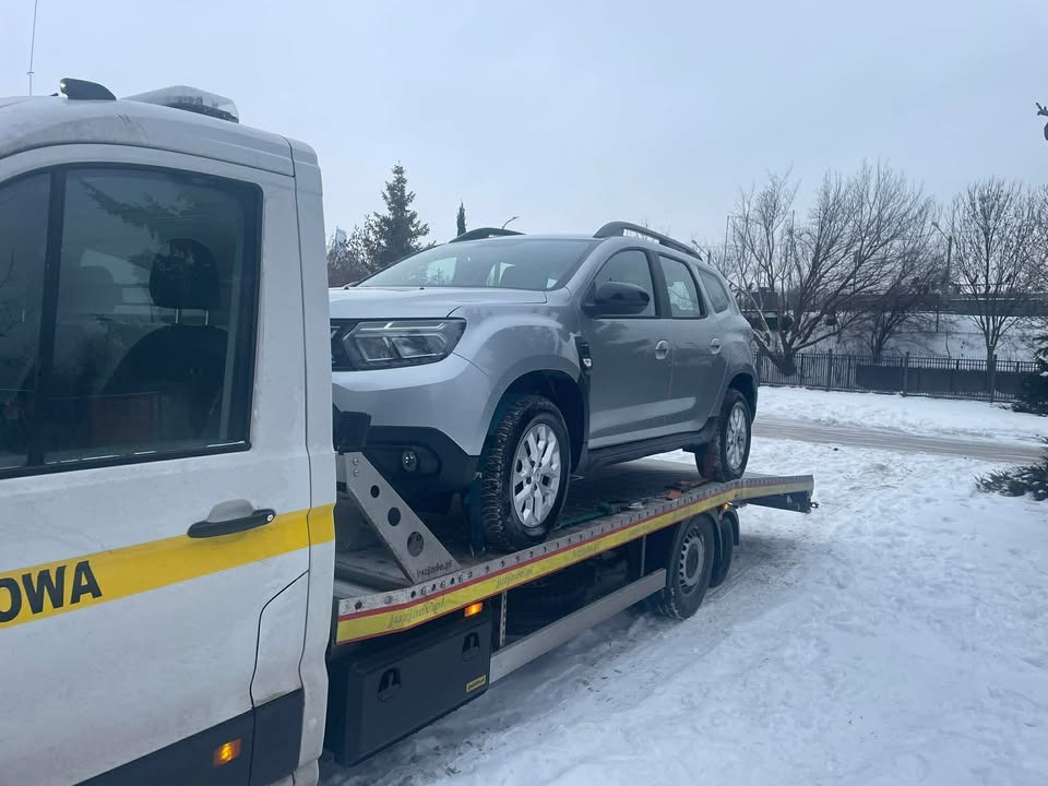 Srebrny Duster na żółtej lawecie w zimowy dzień. Widoczny fragment kabiny lawety z napisem 'OWA'.