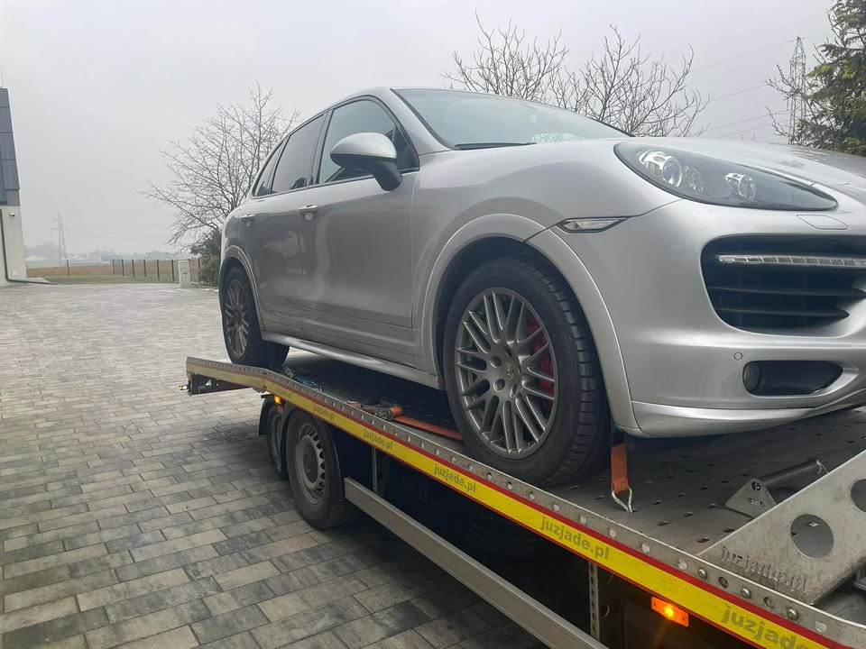 Srebrny Porsche Cayenne na żółtej lawecie z widocznym logo firmy. Auto zabezpieczone pasami transportowymi. Tło: szara kostka brukowa, budynek i drzewa bez liści.