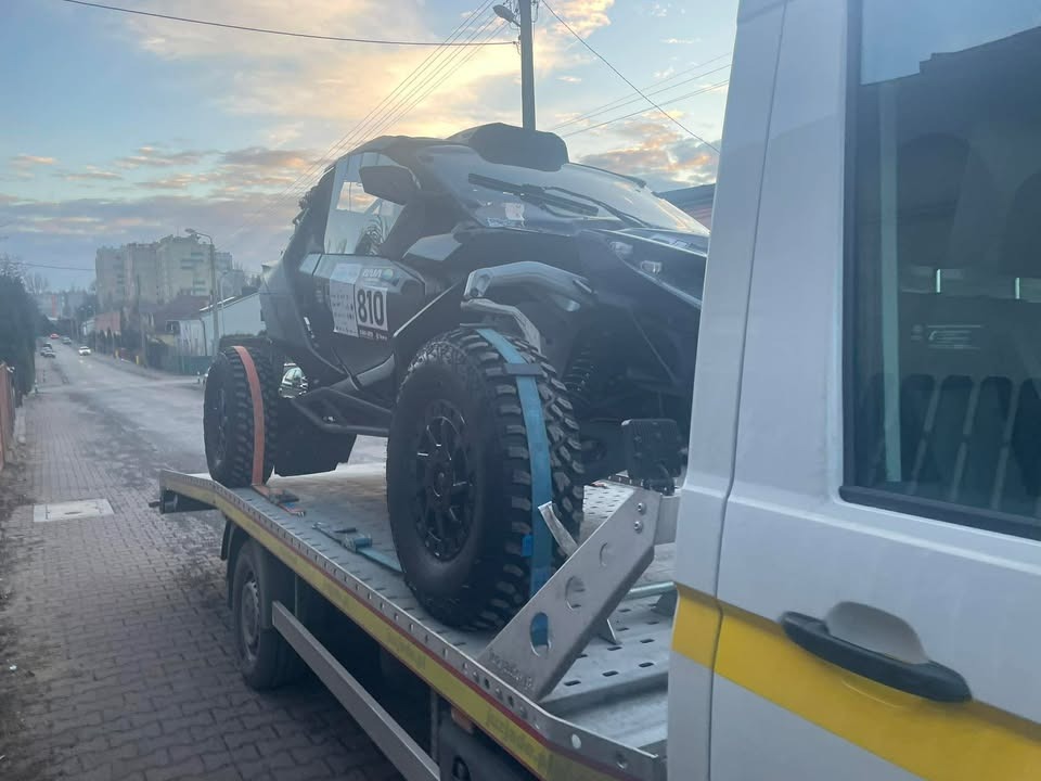 Czarny pojazd terenowy z numerem 810, zabezpieczony pasami, przewożony na platformie lawety na tle miejskiej ulicy w pochmurny dzień.