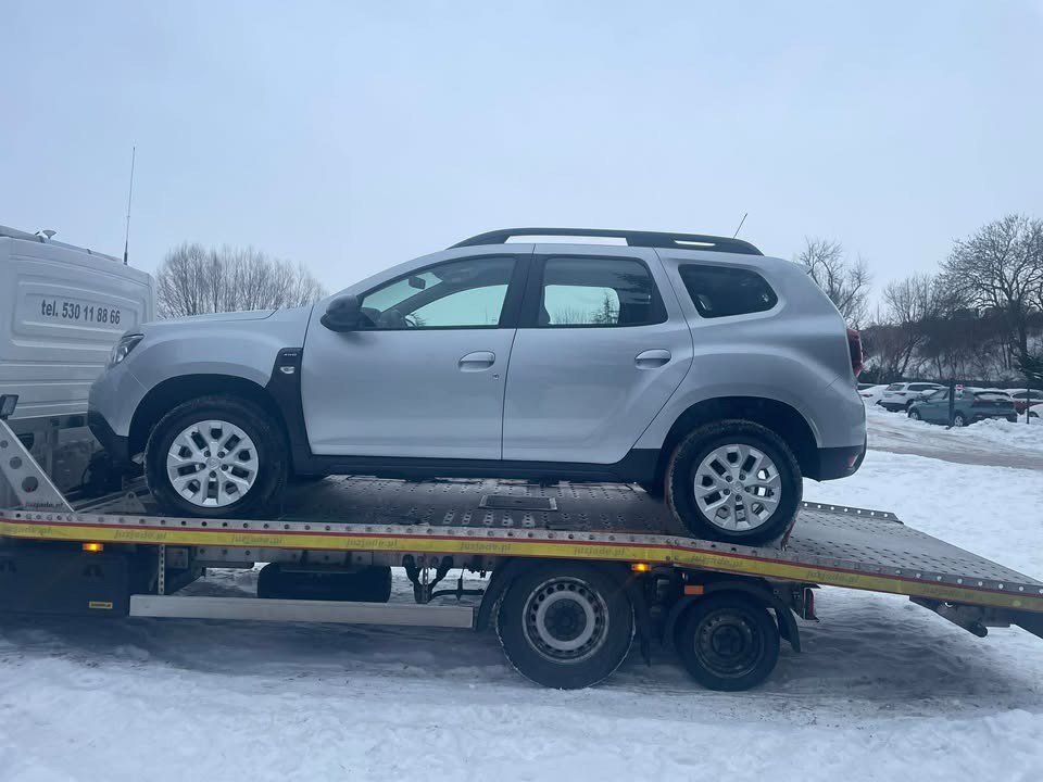 Srebrny SUV na żółtej lawecie w zimowej scenerii. Widoczny numer telefonu na kabinie lawety. Auto zabezpieczone pasami. Tło: parking i drzewa.