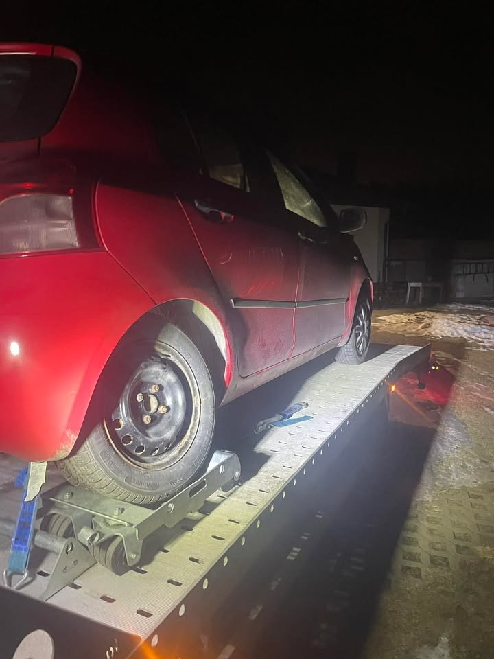 Czerwony samochód osobowy z uszkodzonym kołem, zabezpieczony na platformie lawety w nocnych warunkach. Widoczne detale mocowania i oświetlenie.