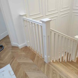 Schody i Stolarstwo - Biała, klasyczna balustrada schodowa z drewna, z dekoracyjnymi panelami ściennymi i parkietem w jodełkę, Radzymin. Detal wykończenia wnętrza.