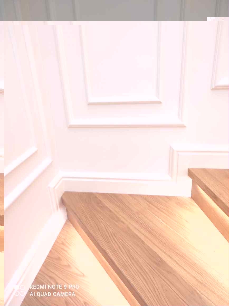 Jasne, drewniane stopnie schodów z subtelnym oświetleniem LED, kontrastujące z białą, klasyczną boazerią. Ujęcie z dołu, podkreślające detale wykończenia.