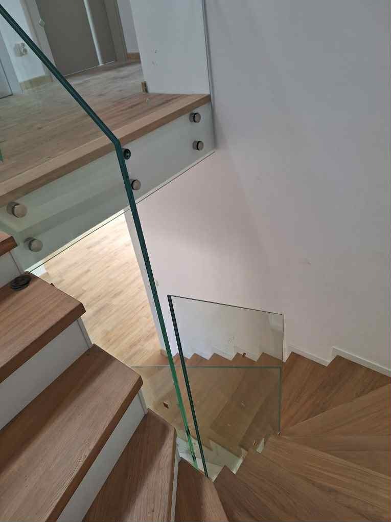 Nowoczesne schody z drewna i szkła, widok z góry. Jasne drewno stopni kontrastuje z minimalistyczną, szklaną balustradą. Elementy mocujące balustradę są srebrne.