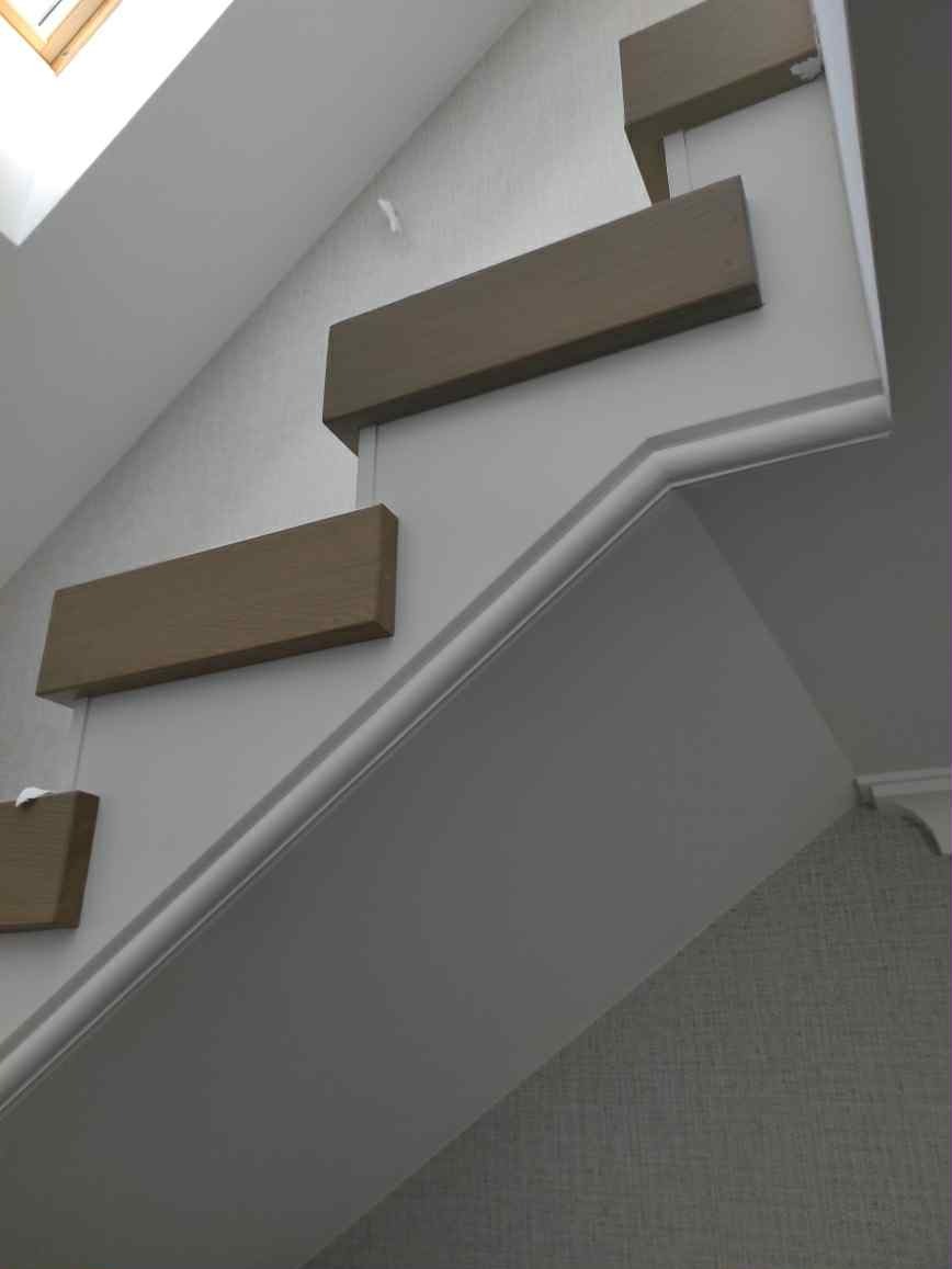 Nowoczesne schody wspornikowe z drewnianymi stopniami w odcieniach brązu, zamontowane przy ścianie z tapetą o delikatnej fakturze. Widok z dołu, oświetlenie naturalne.