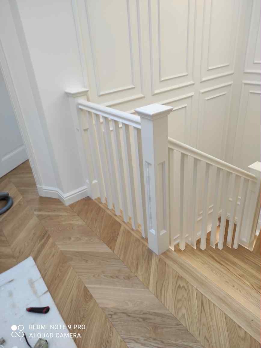 Biała, klasyczna balustrada schodowa z drewna, z dekoracyjnymi panelami ściennymi i parkietem w jodełkę, Radzymin. Detal wykończenia wnętrza.