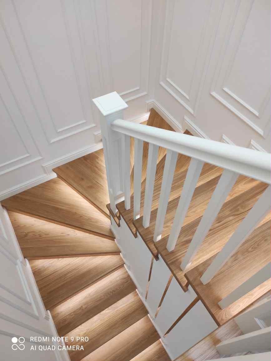 Kaskadowe schody z jasnego drewna z białymi balustradami, widok z góry. Stopnie podświetlone, ściany z dekoracyjnymi panelami. Nowoczesny design.