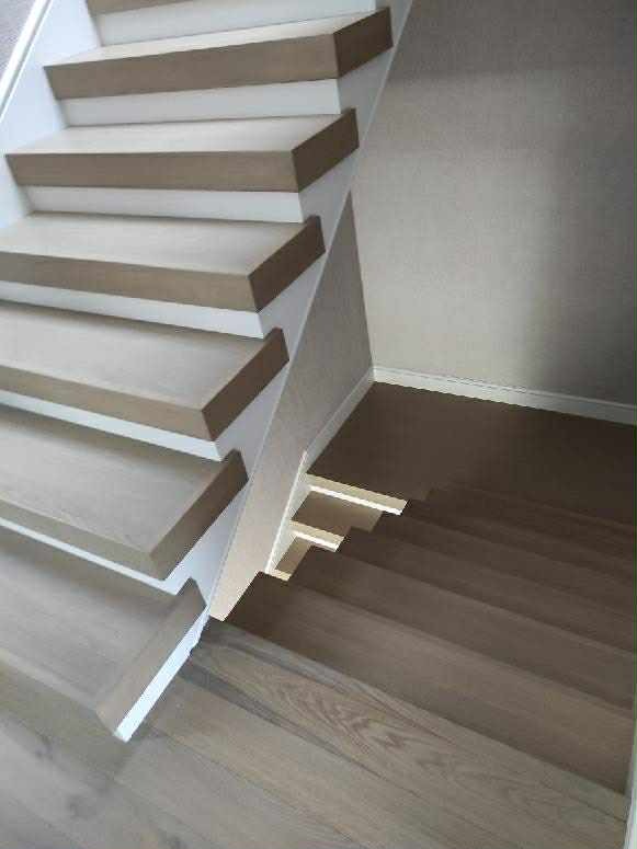 Jasne, nowoczesne schody z drewnianymi stopniami i białymi podstopnicami, widok z góry, ukazujący detale wykończenia i subtelne oświetlenie pod schodami.