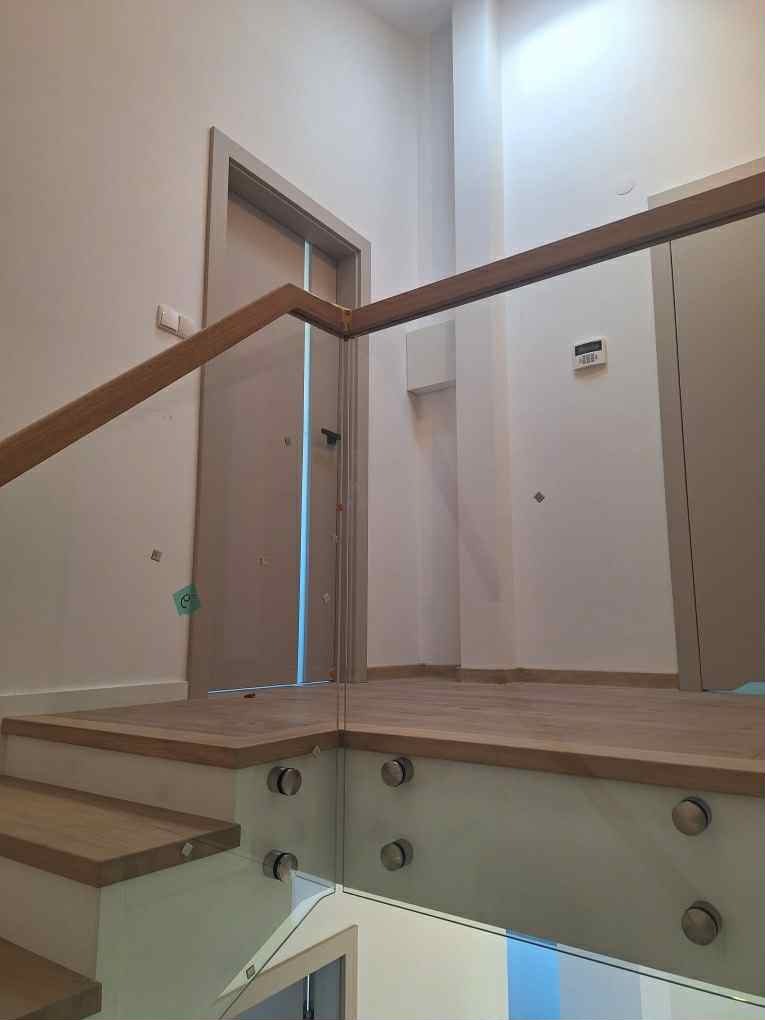 Nowoczesne schody z drewnianymi stopniami i szklaną balustradą mocowaną na metalowych uchwytach. Widoczny fragment drzwi i jasne ściany w tle.