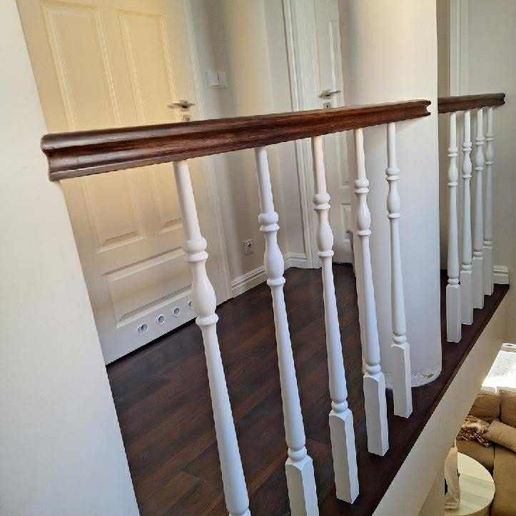 Białe, toczone słupki balustrady z ciemnobrązowym poręczą, widoczne na tle ciemnej podłogi i białych drzwi, ujęcie z góry.
