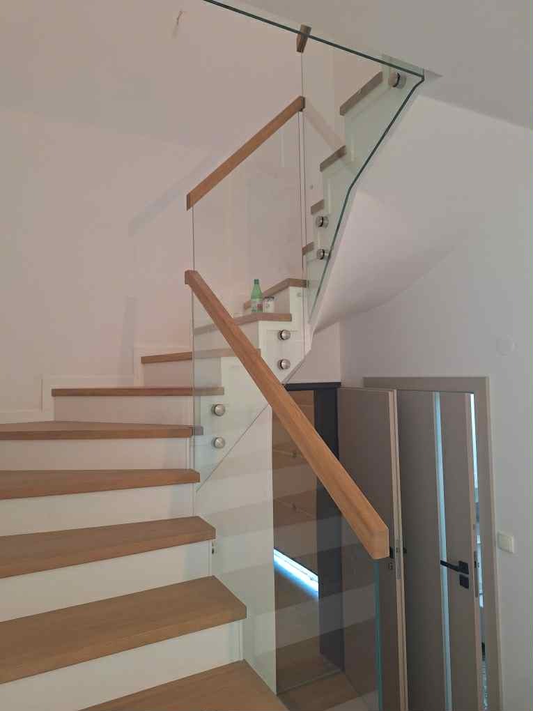 Nowoczesne schody z drewnianymi stopniami i szklaną balustradą, mocowaną na metalowych uchwytach. Widok z dołu, z fragmentem drzwi w tle.