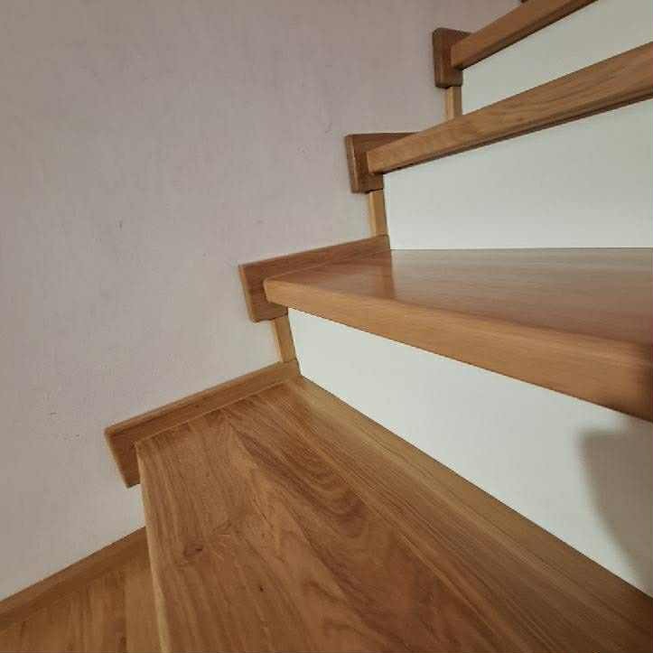 Jasne, dębowe stopnie schodów z białymi podstopnicami, widziane z dołu pod kątem. Widoczne usłojenie drewna i fragment ściany w tle. Minimalistyczny design.