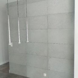 PORTAL Maciej Hyżowiecki - Ściana wyłożona szarymi, prostokątnymi płytami imitującymi beton, oświetlona trzema wiszącymi lampami z metalowymi, smukłymi kloszami. Minimalistyczny design.