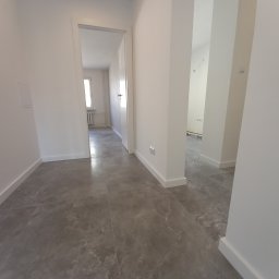 PORTAL Maciej Hyżowiecki - Jasne wnętrze po remoncie: szare płytki podłogowe imitujące kamień, białe ściany i listwy przypodłogowe. Widok na korytarz i pokoje przez otwarte drzwi.