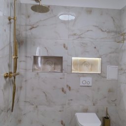 PORTAL Maciej Hyżowiecki - Wykończona łazienka z marmurowymi płytkami, złotą armaturą i podświetlanymi niszami z motywem kwiatowym. Widoczny prysznic, toaleta podwieszana i akcesoria.
