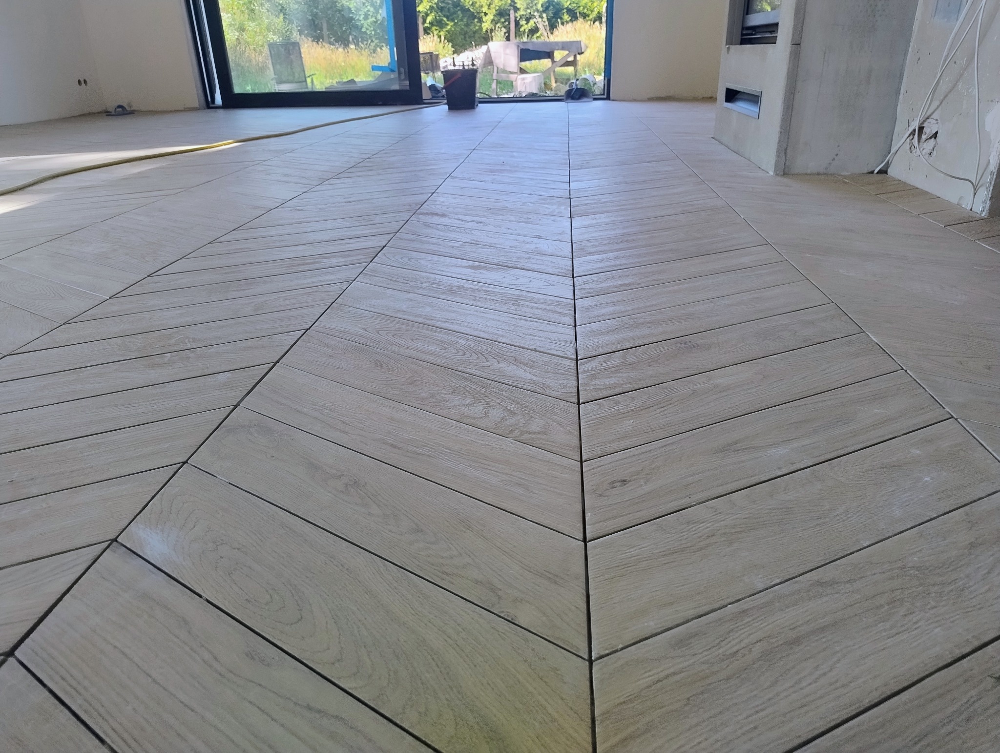 Ułożona pod kątem jasna podłoga z płytek imitujących drewno, widok z perspektywy podłogi, z oknem w tle. Widać fragment ściany z instalacją elektryczną.