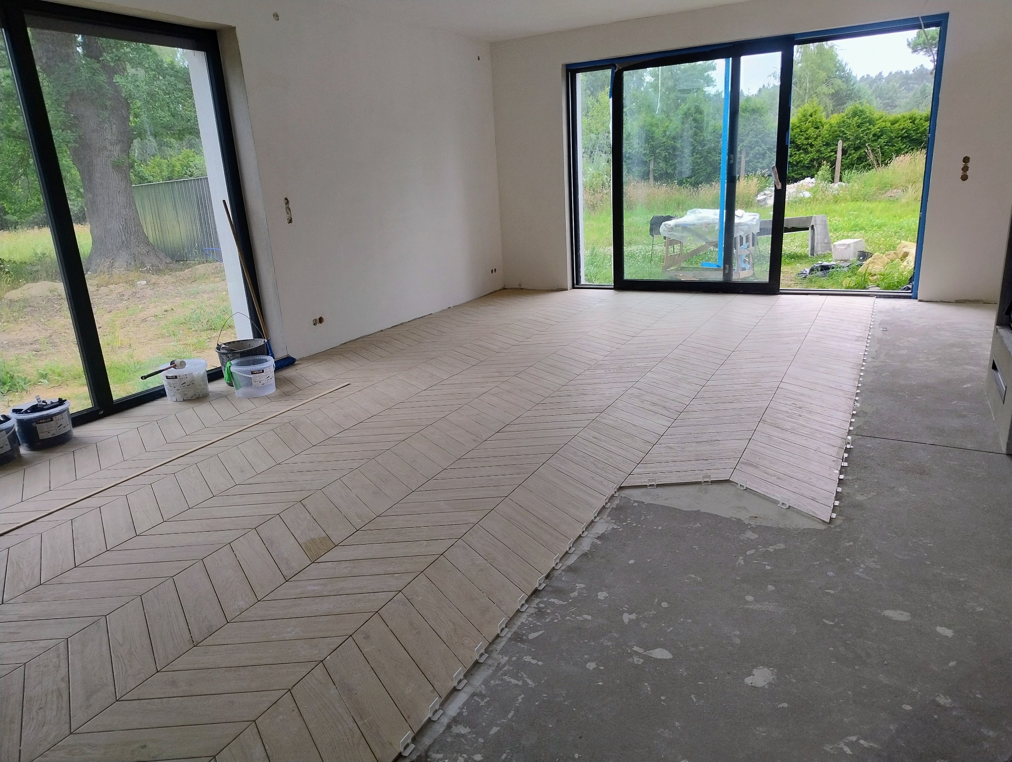 Jasne płytki ułożone we wzór jodełki na podłodze w przestronnym wnętrzu z dużymi oknami. Część podłogi jeszcze nieukończona, widoczne narzędzia i materiały.