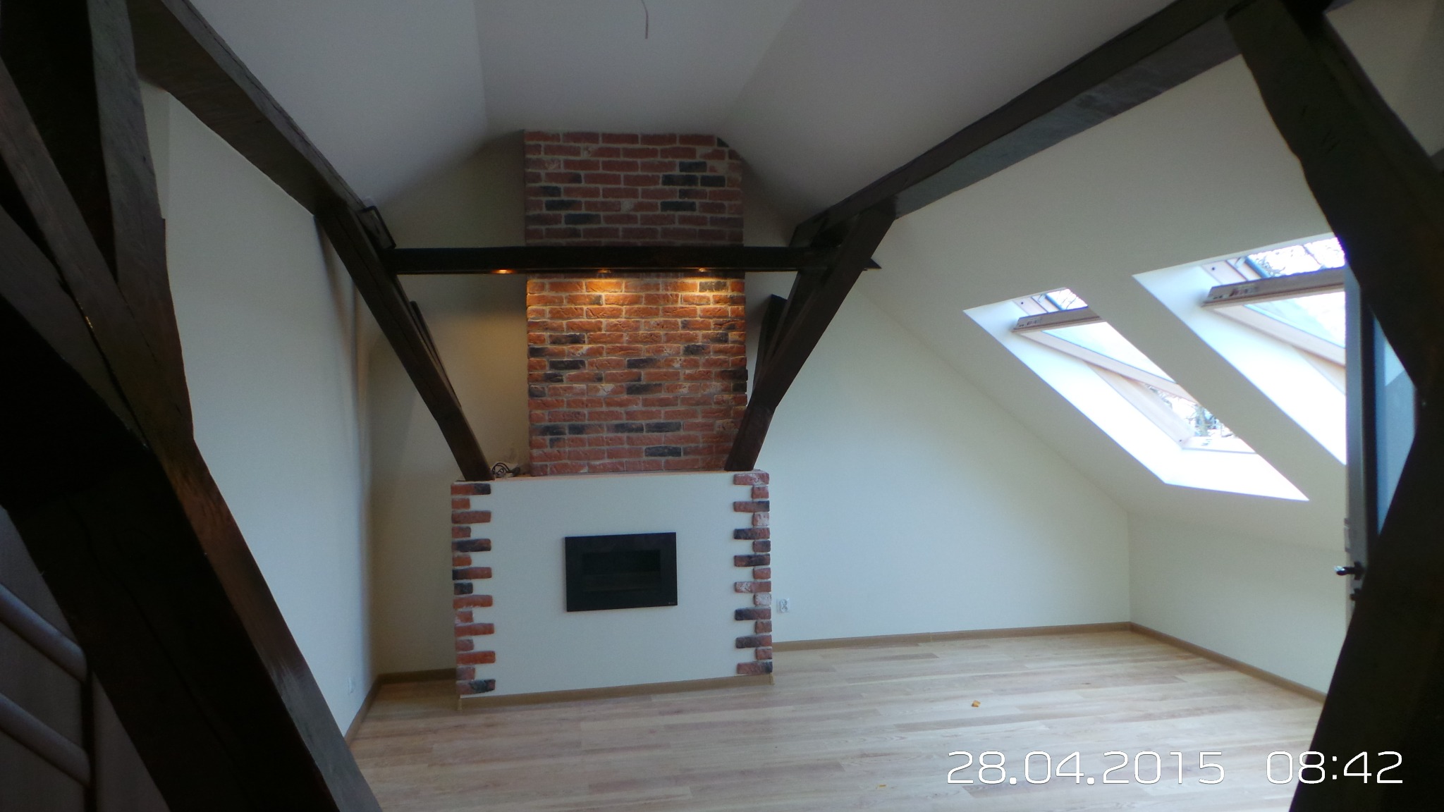 Poddasze po remoncie: drewniane belki, ceglany kominek z oświetleniem, jasne ściany i podłoga, okna dachowe. Widok z dołu, z fragmentem schodów.