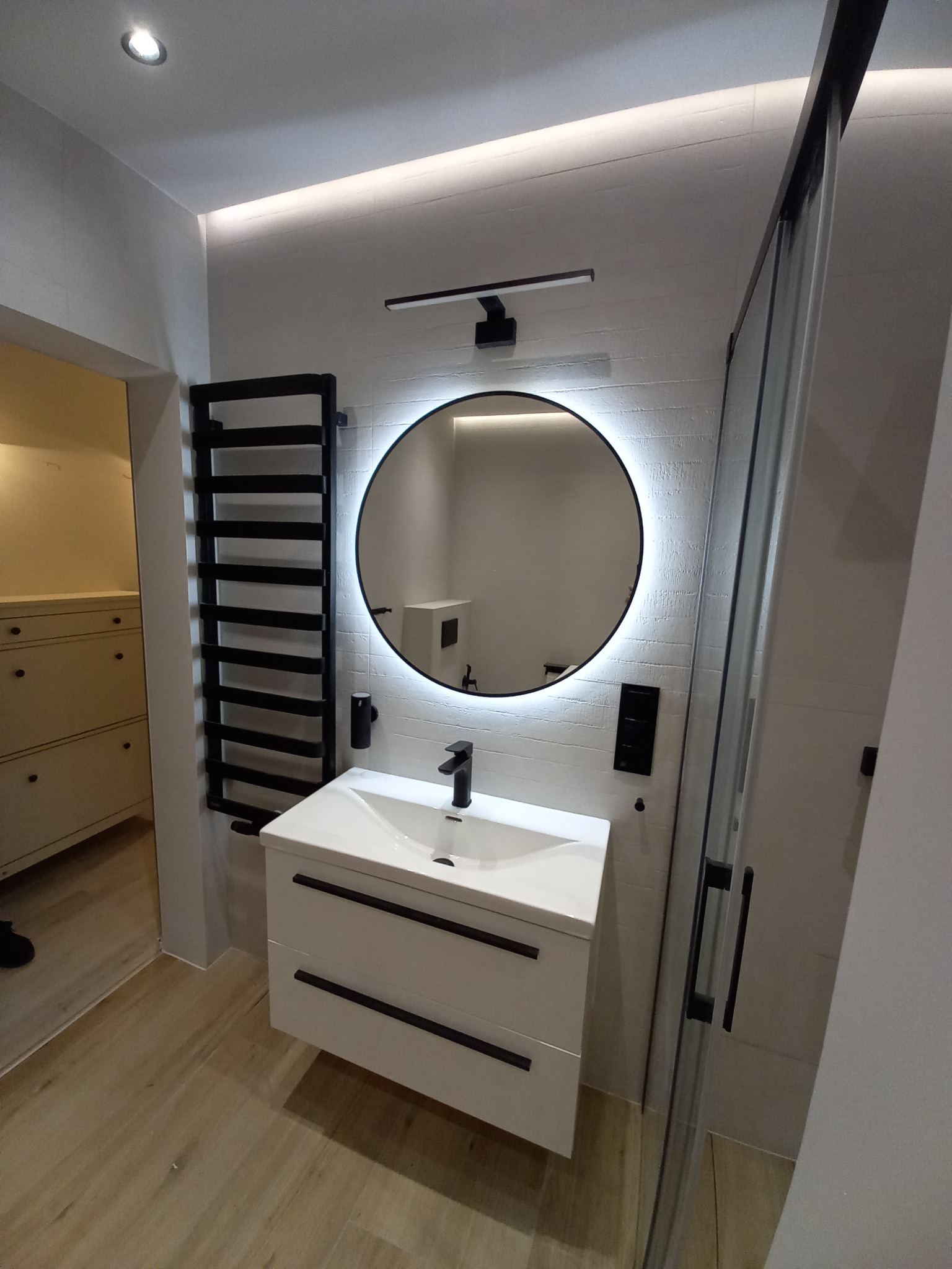 Nowoczesna łazienka z okrągłym lustrem LED, białą szafką z czarnymi uchwytami i grzejnikiem drabinkowym. Jasne płytki na ścianach i podłodze.