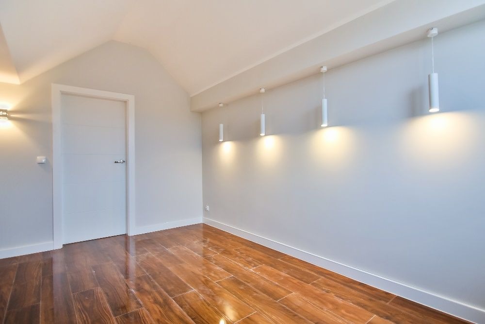 Jasne wnętrze po remoncie: parkiet w odcieniach brązu, białe ściany z oświetleniem punktowym i białe drzwi. Poddasze z delikatnym skosem.