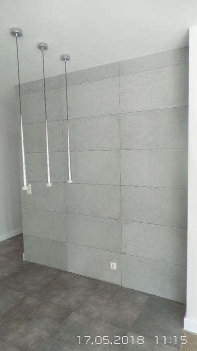 Ściana wyłożona szarymi, prostokątnymi płytami imitującymi beton, oświetlona trzema wiszącymi lampami z metalowymi, smukłymi kloszami. Minimalistyczny design.
