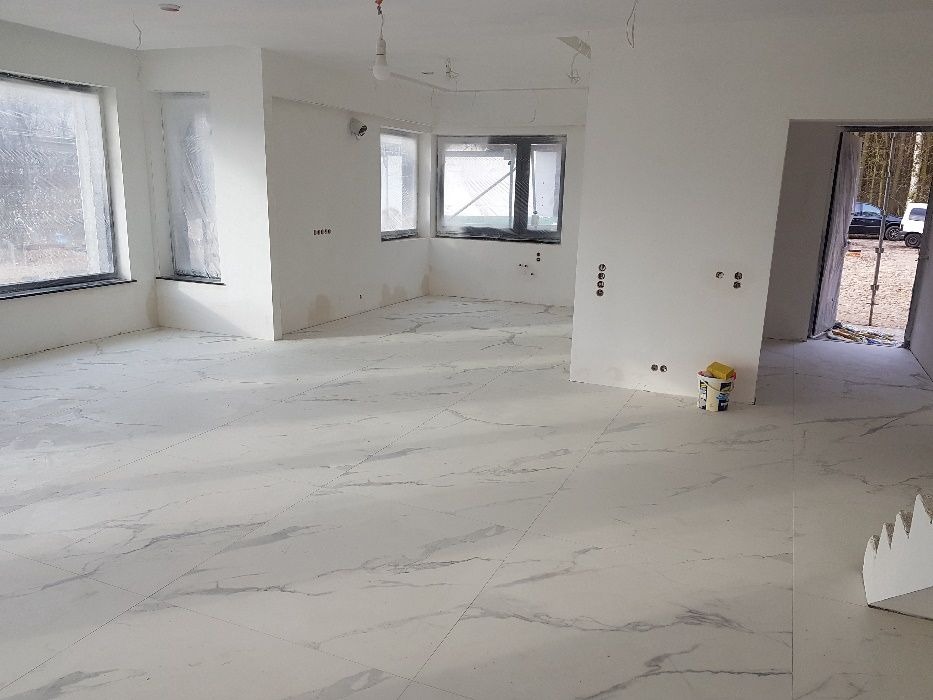 Jasne, marmurowe płytki podłogowe w przestronnym, surowym wnętrzu. Okna zabezpieczone folią, ściany przygotowane do malowania, widoczne puszki elektryczne.