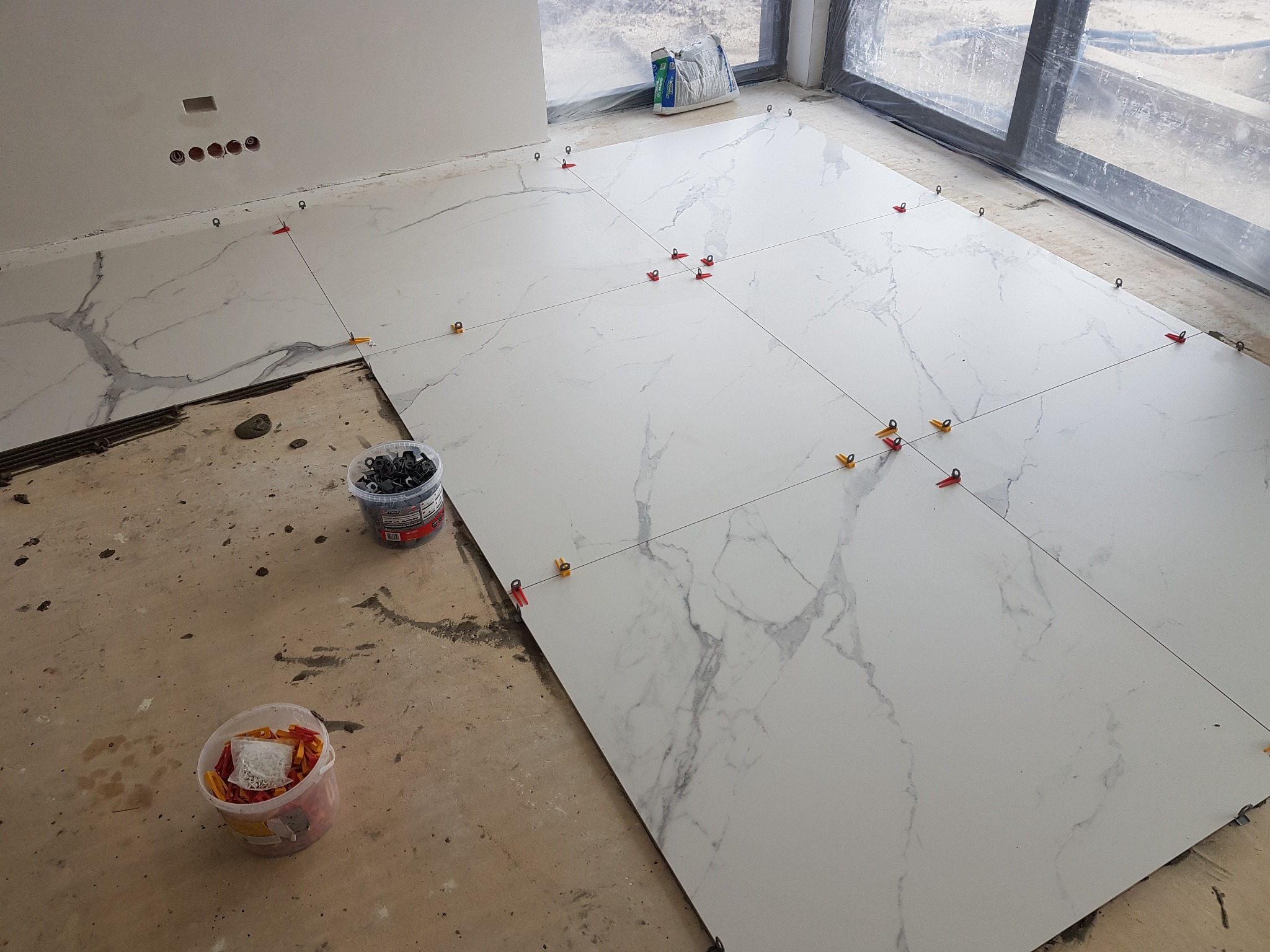 Białe płytki imitujące marmur, ułożone na podłodze z użyciem klipsów poziomujących. Częściowo wykończona podłoga, widoczne narzędzia i materiały budowlane.