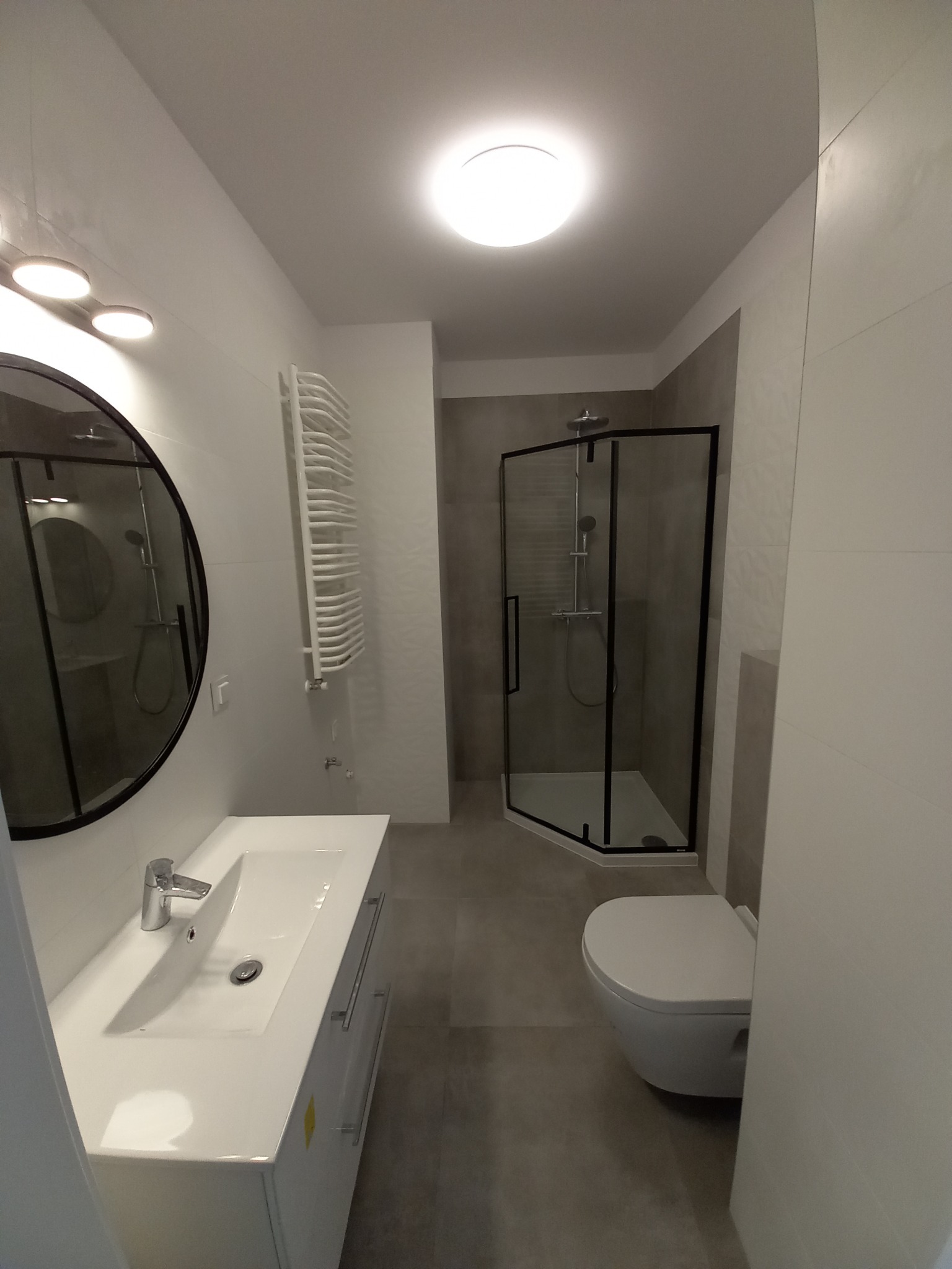 Nowoczesna łazienka w szaro-białej tonacji z kabiną prysznicową z czarnymi ramami, owalnym lustrem i grzejnikiem drabinkowym. Minimalistyczny design.