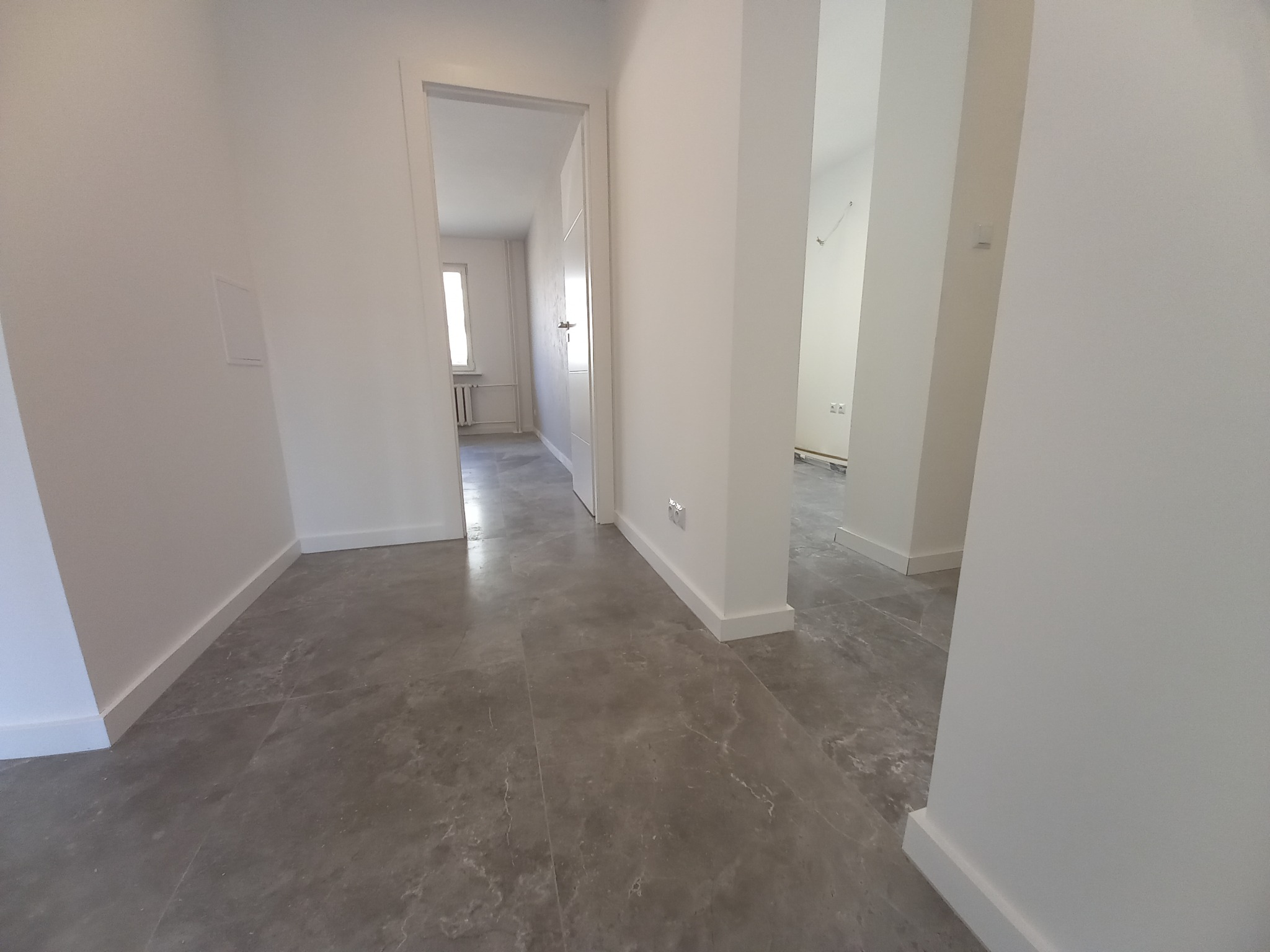 Jasne wnętrze po remoncie: szare płytki podłogowe imitujące kamień, białe ściany i listwy przypodłogowe. Widok na korytarz i pokoje przez otwarte drzwi.