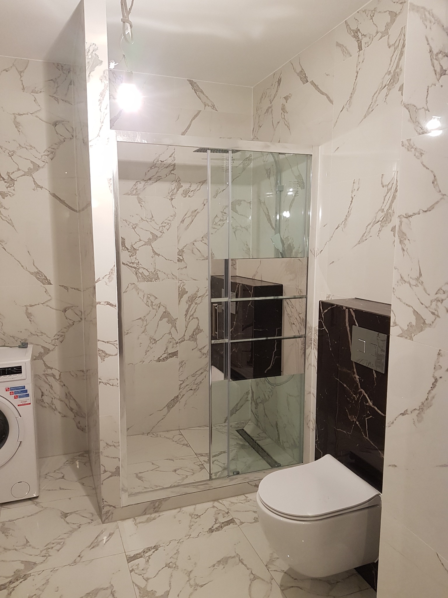 Wykończona łazienka z kabiną prysznicową, toaletą podwieszaną i pralką. Płytki imitujące marmur na ścianach i podłodze. Nowoczesny design.
