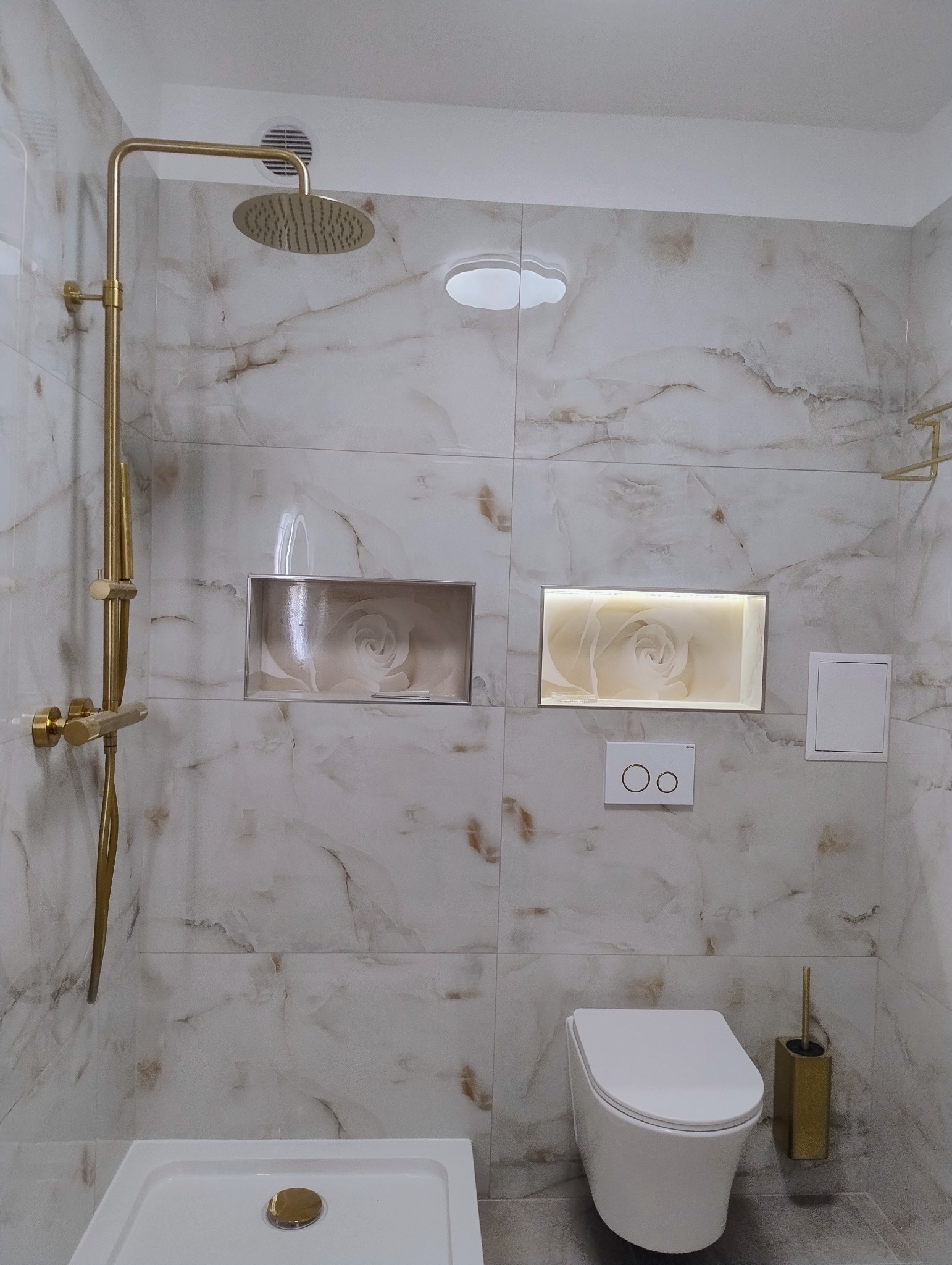 Wykończona łazienka z marmurowymi płytkami, złotą armaturą i podświetlanymi niszami z motywem kwiatowym. Widoczny prysznic, toaleta podwieszana i akcesoria.